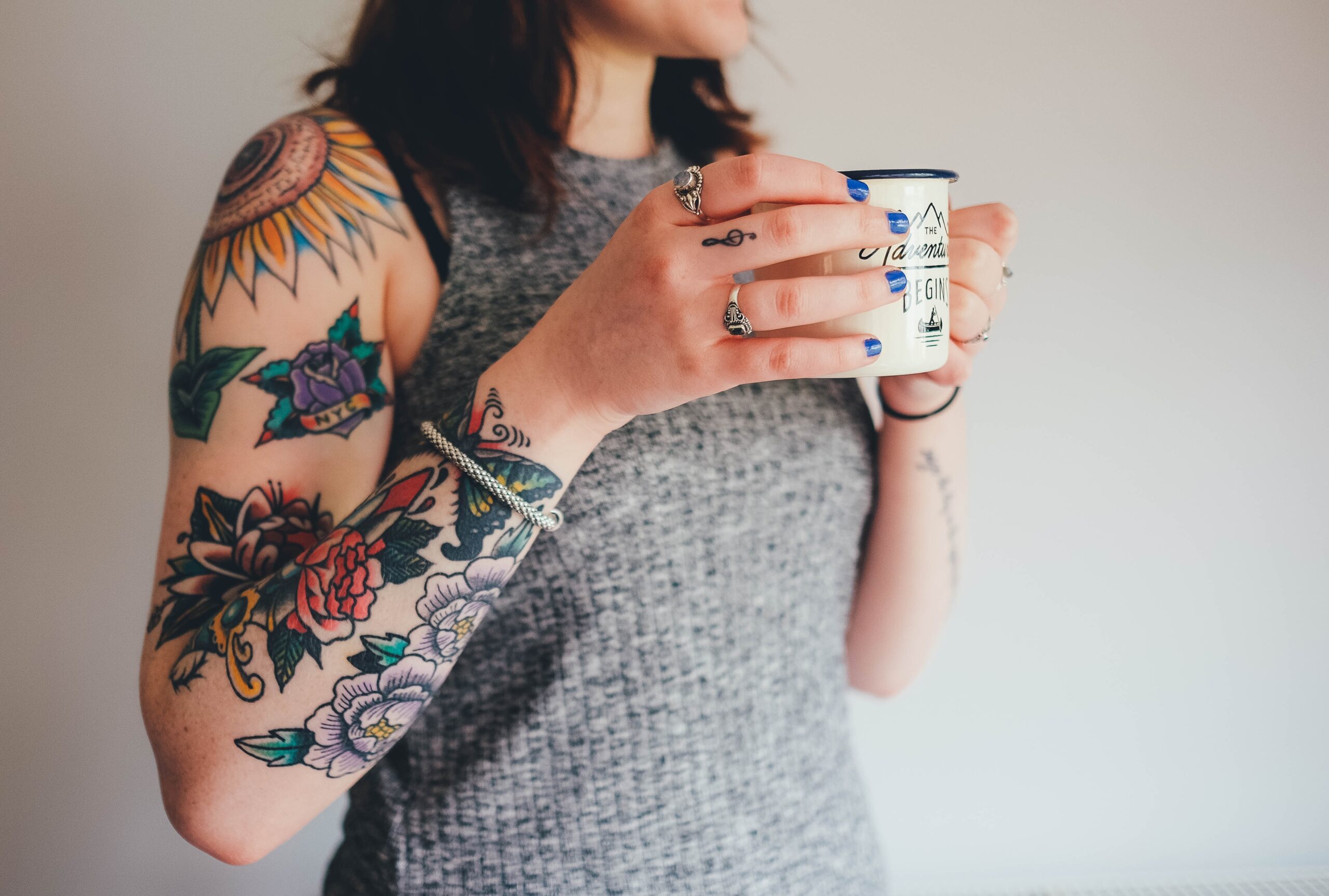 A woman in a tank top with a sleeve of tattoos drinking coffee in front of a grey wall.